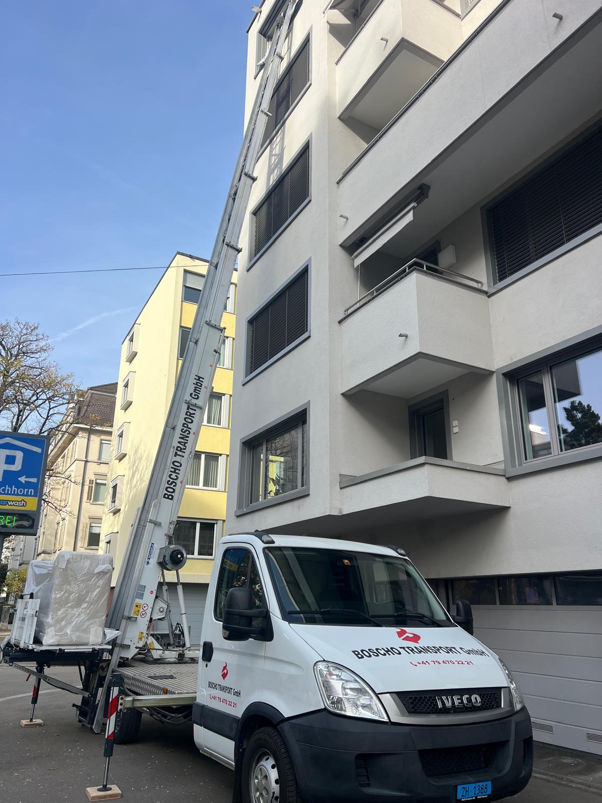 White utility truck with a tall hydraulic crane extending beside a modern apartment building, lifting or positioning materials on a street-side construction site.